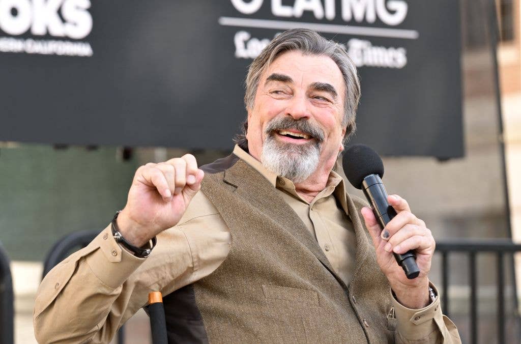 Tom Sellleck sitting on a stage, holding a microphone, and smiling