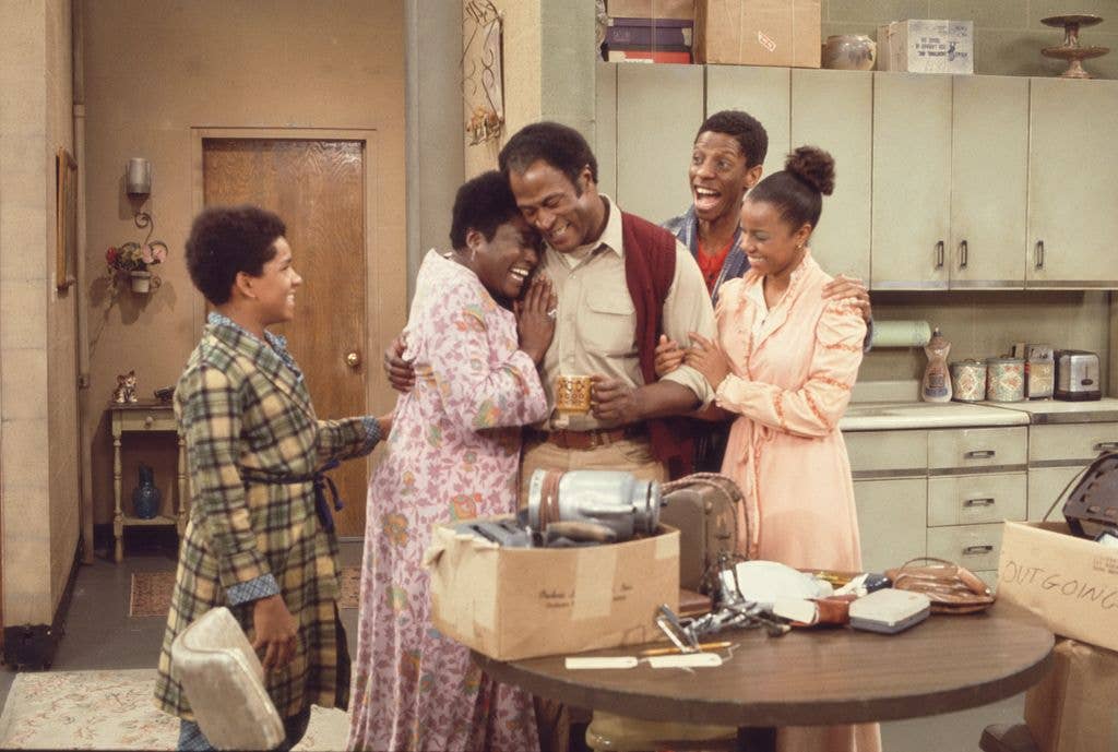 A group of five people share a laughing group hug around a father figure in a kitchen filled with packed moving boxes.