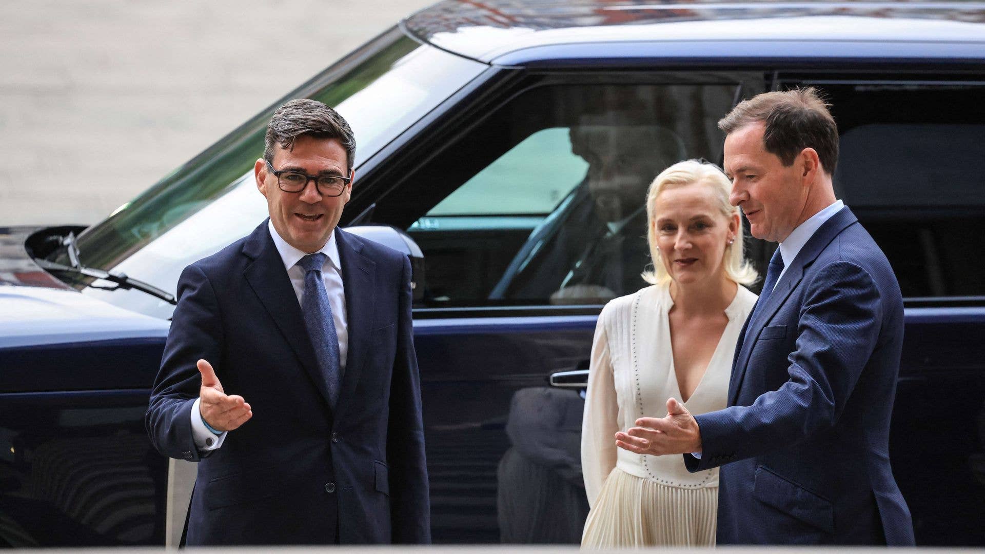 Two men in suits and a woman in a white dress stand by a car. The man on the left wears glasses.