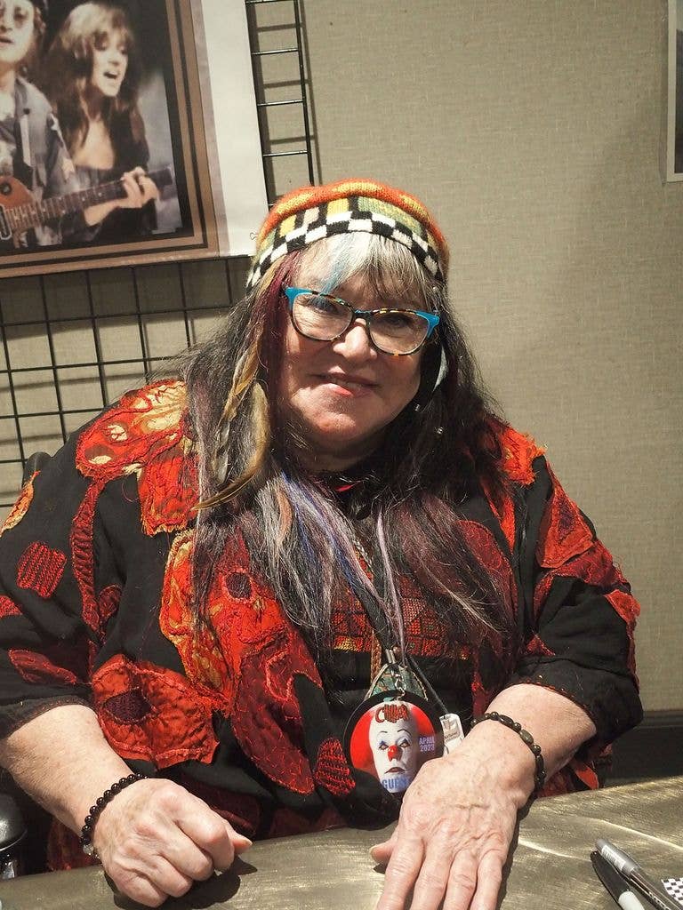 Melanie Safka, wearing a colourful knitted beanie and a patchwork jacket, sits at a signing table with a Chiller Theatre guest badge around her neck.