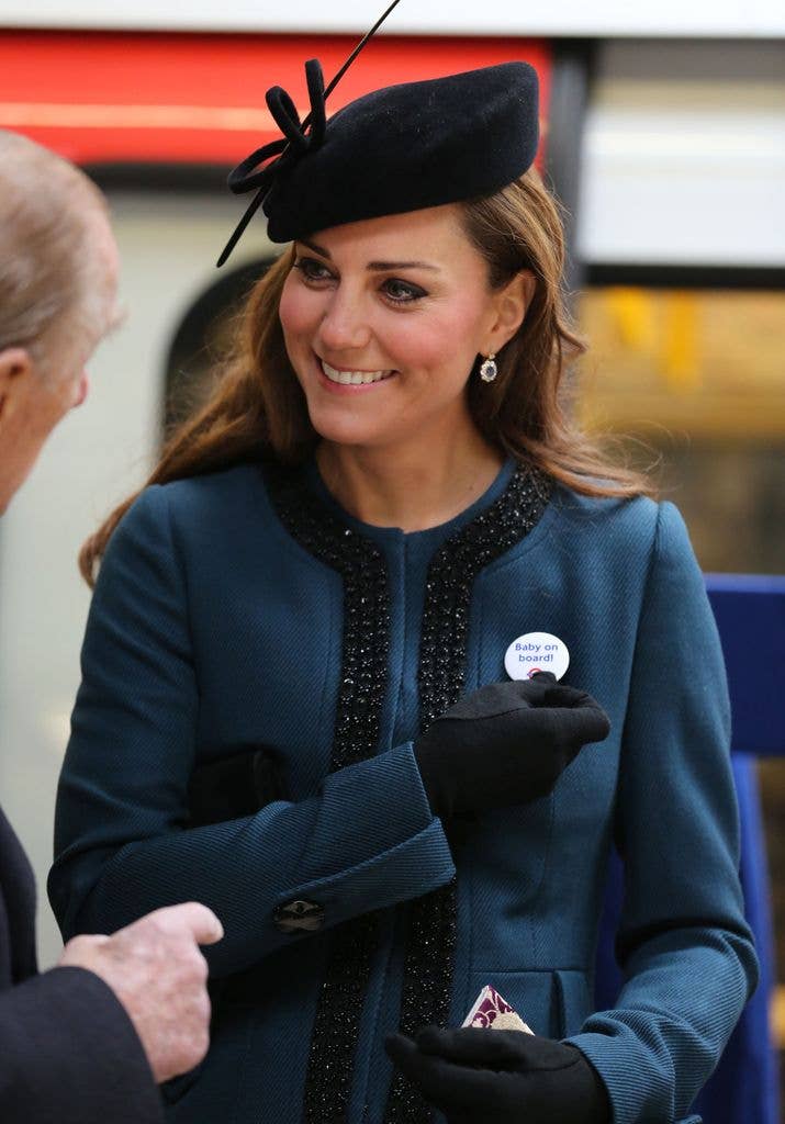 Kate Middleton, wearing a blue coat and black hat, smiles and talks to another person.