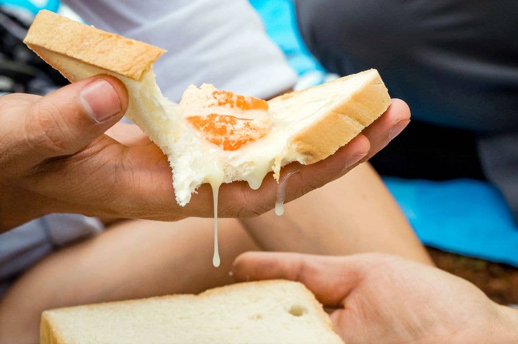 A hand holds a torn piece of bread topped with tangerine segments and dripping condensed milk, with another slice of bread visible below.