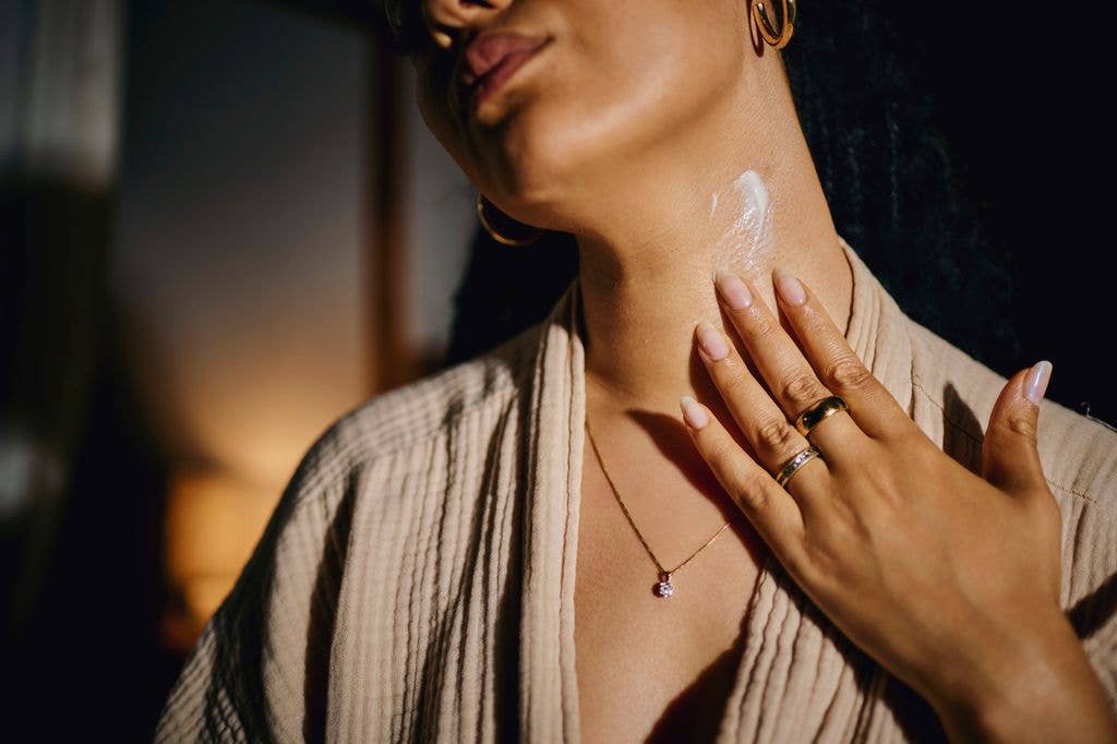 Young adult Black woman wearing beige textured robe applies moisturizing lotion to her neck in a softly lit indoor setting, focusing on gentle morning self care and skincare routine