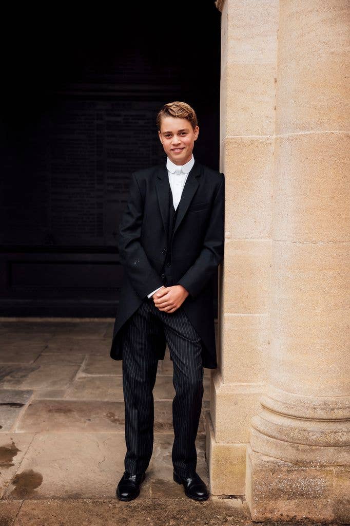 Prince George of Cambridge stands by a stone pillar, wearing a black tuxedo with a white shirt and black shoes.