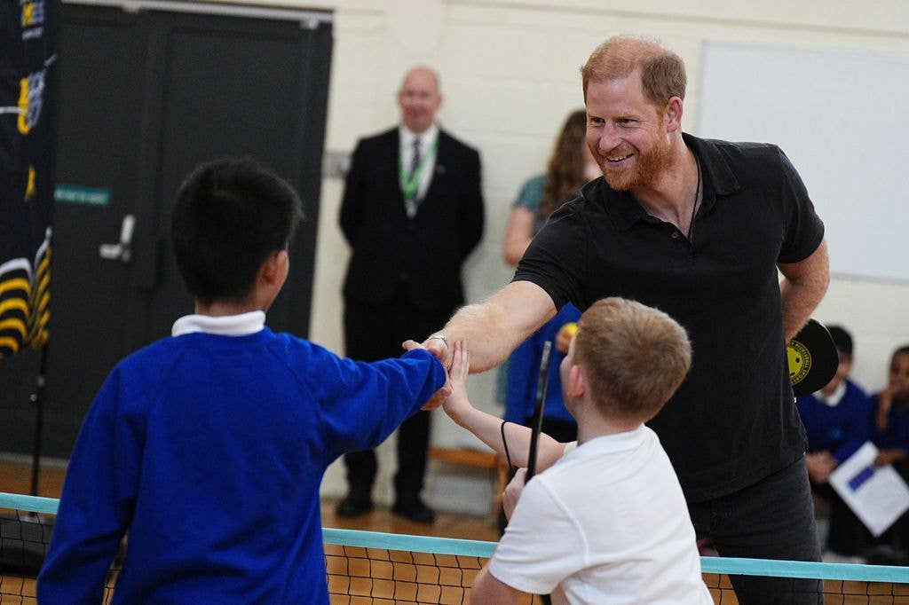 Prince Harry, wearing a black polo shirt, smiles and shakes hands over a net with schoolchildren during a sports session in a gym hall.