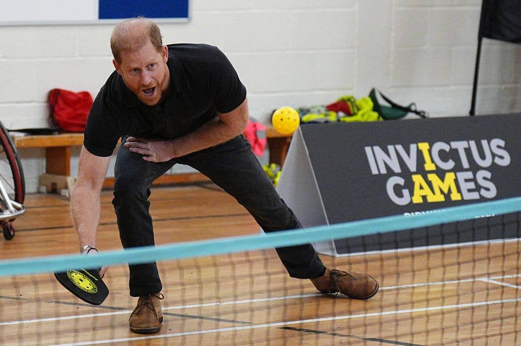 Prince Harry, in a black polo shirt, leans low with a paddle to return a shot on a pickleball court, with an Invictus Games sign visible behind him.