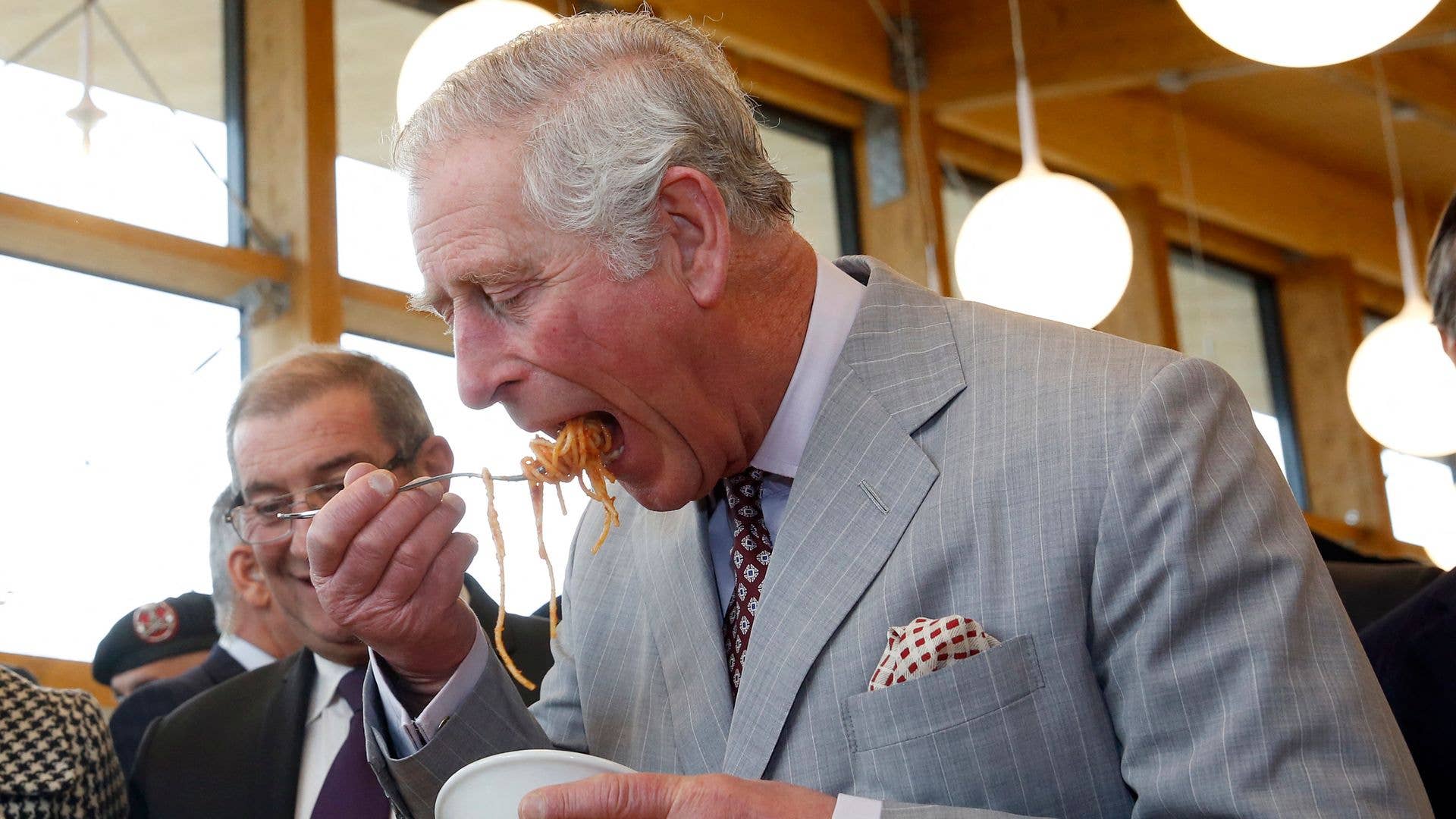 King Charles in a gray suit, eating pasta with a spoon.