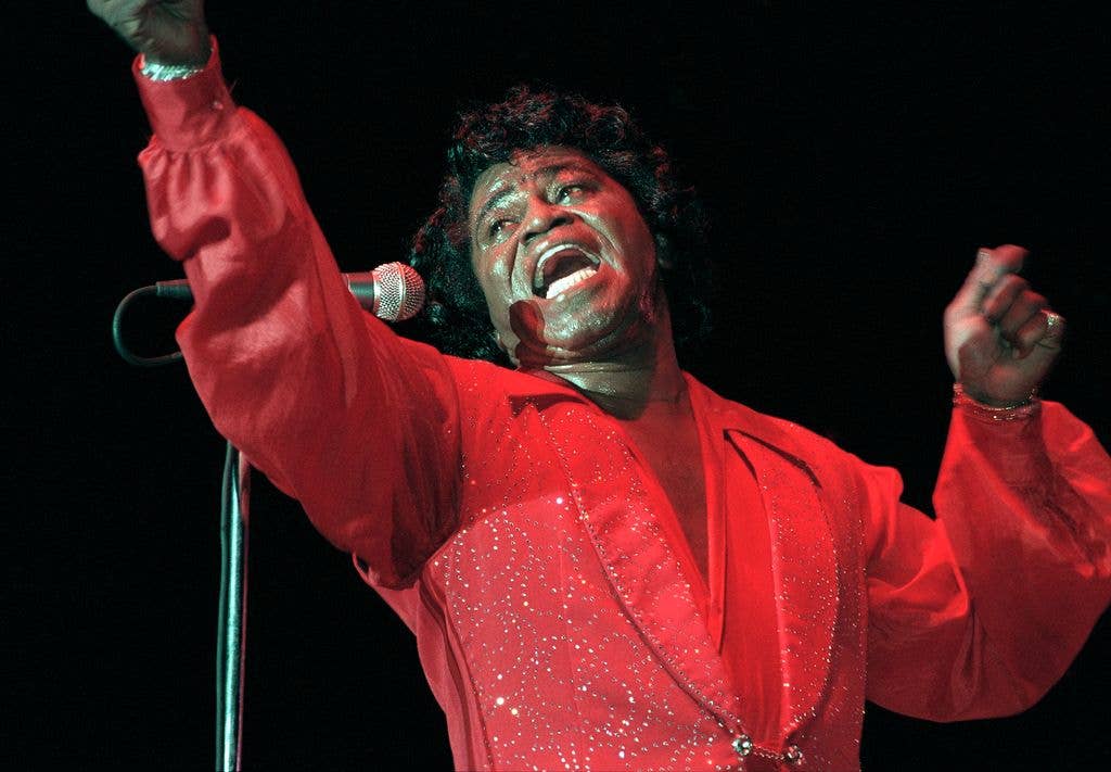 James Brown, dressed in a sparkling red suit, sings passionately into a microphone with his fist raised on stage.