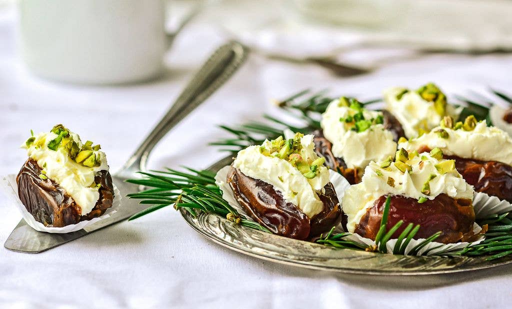 Several dates filled with whipped cream cheese and sprinkled with chopped pistachios are arranged on a silver plate garnished with pine sprigs.