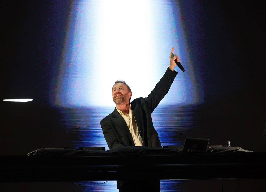 David Guetta raises a microphone above his head while performing on stage behind a DJ booth, with a bright light illuminating a large screen behind him.