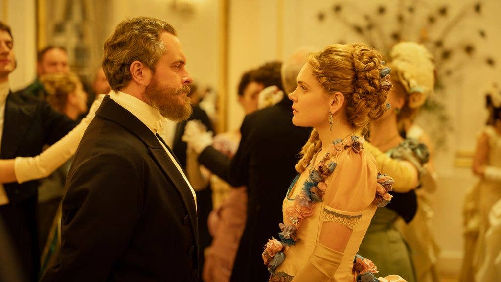 Tomasz Schuchardt in a black suit and bow tie looks at a woman in a yellow gown with floral details.