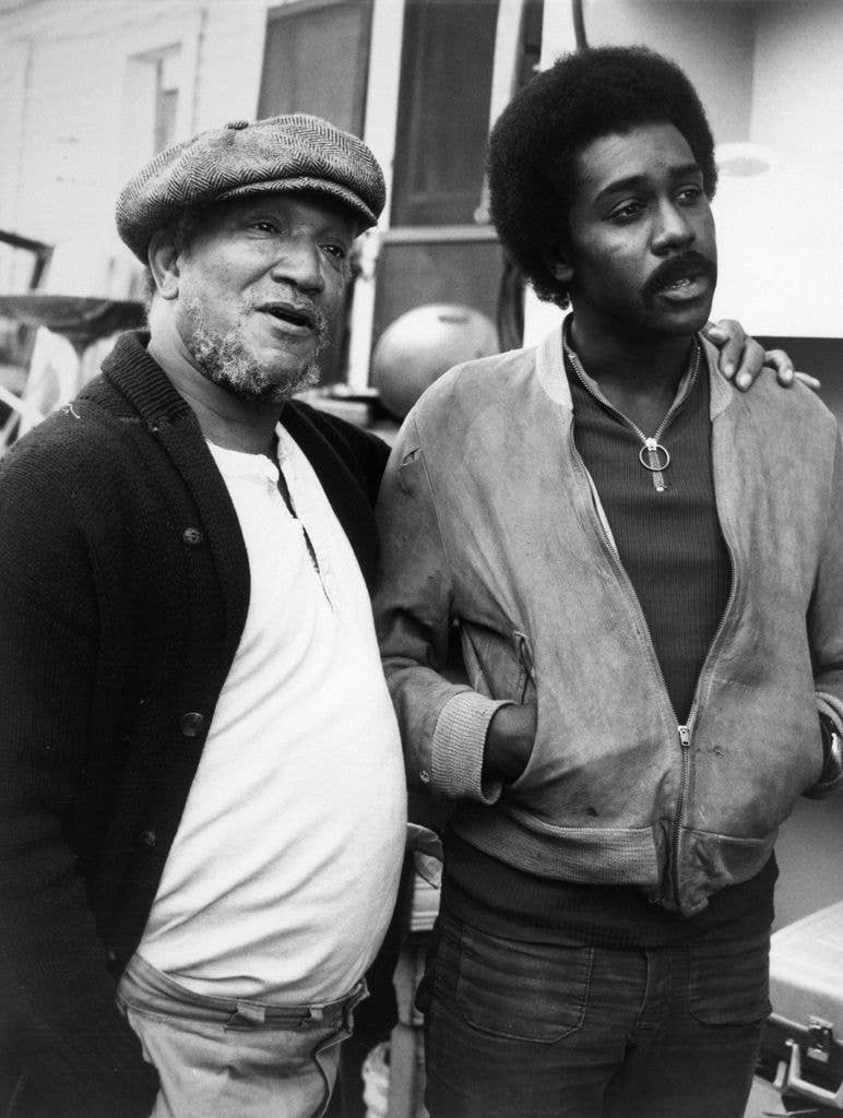 Black and white photo of Redd Foxx, wearing a flat cap and cardigan, standing beside Demond Wilson, who has his arm around him, both looking off to the side.