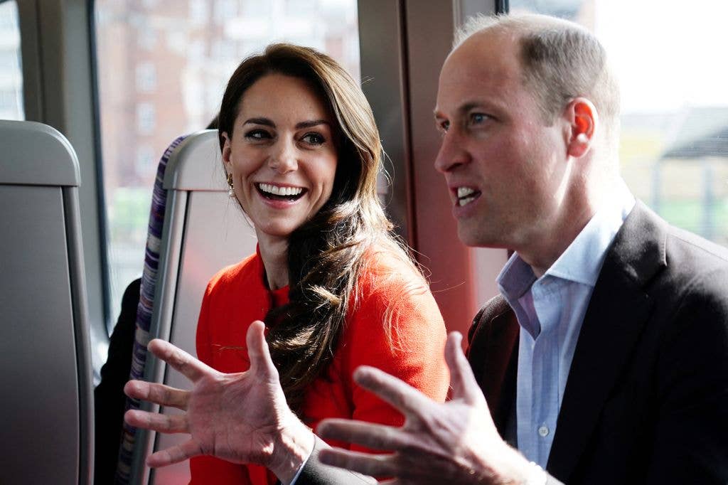 Prince William and Kate Middleton smiling and gesturing while seated on a train.