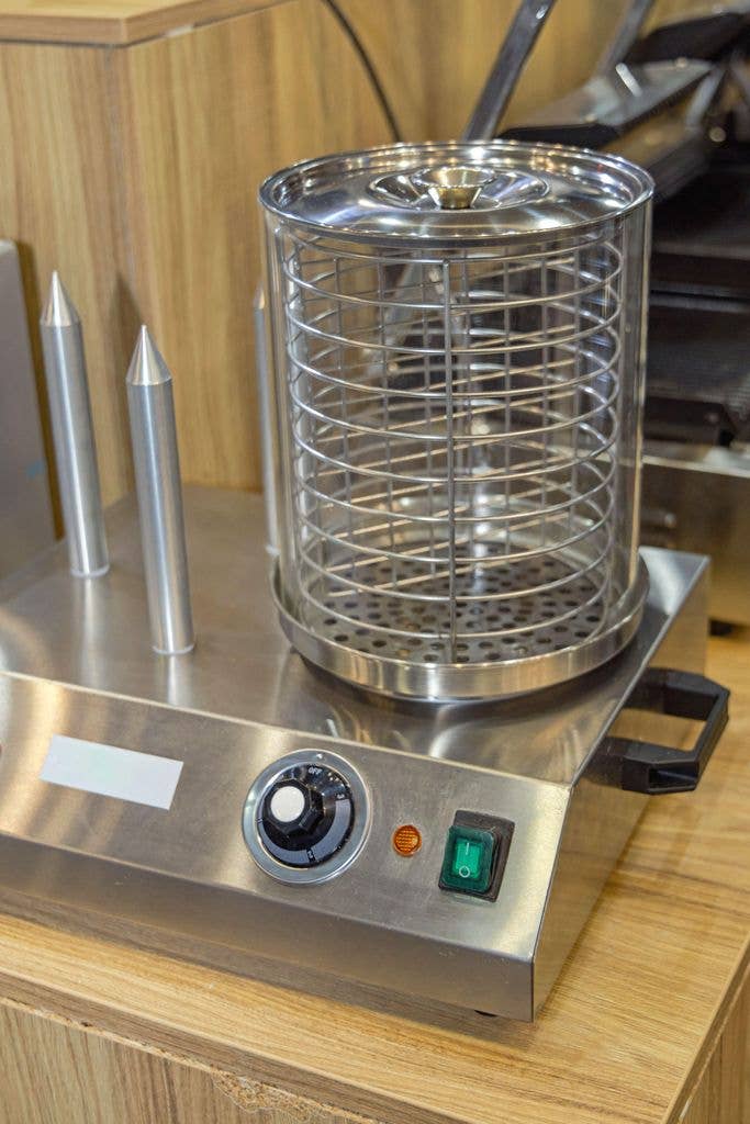 A cylindrical stainless steel hot dog steamer with a wire basket and metal bun-warming cones stands on a wooden counter, its control panel showing a dial, indicator light and green power switch.