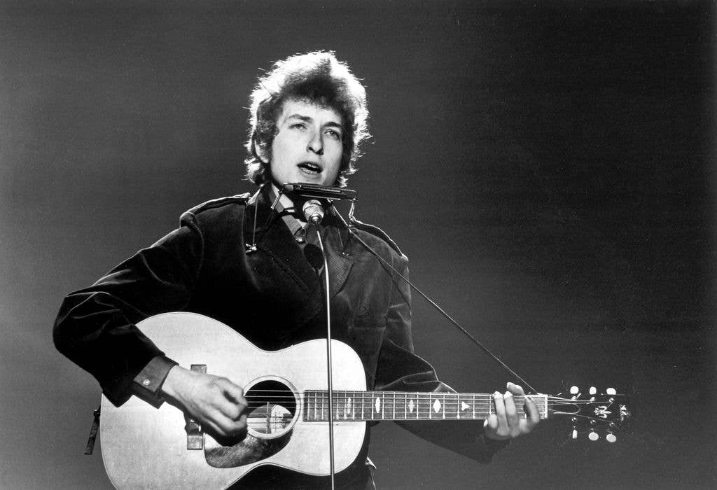 Black and white photograph of Bob Dylan singing into a microphone with a harmonica rack around his neck while playing an acoustic guitar.