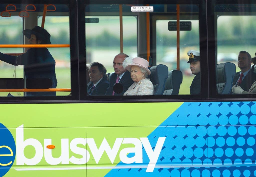 Queen Elizabeth II in a pink hat sits on a busway bus with other individuals.