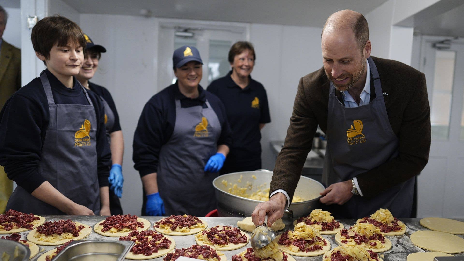 Prince William in a brown suit and apron, adds toppings to pies in a kitchen with staff.