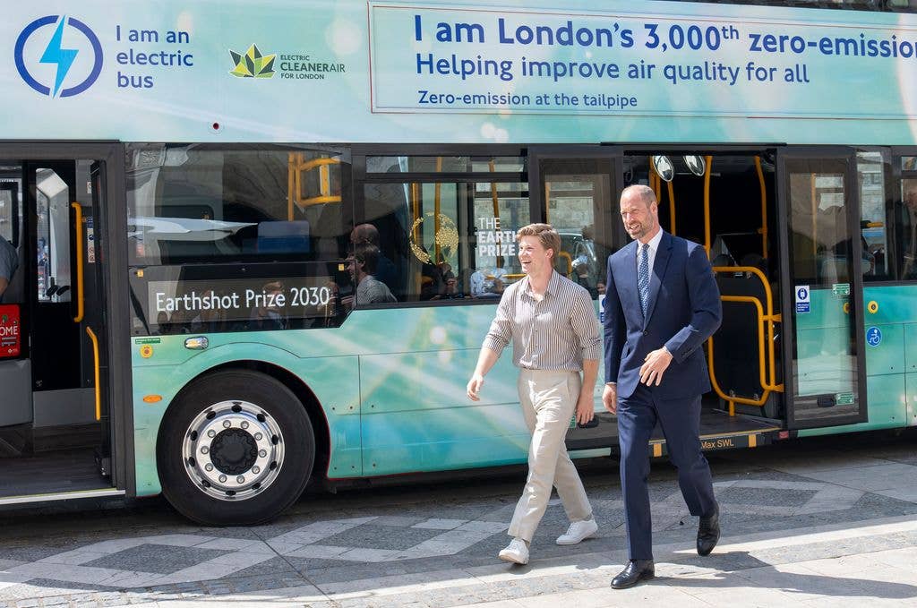 Prince William in a blue suit, walking with Robert Irwin, wearing a striped shirt and khaki pants, beside a zero-emission bus.