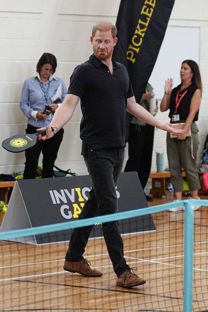 Prince Harry, holding a pickleball paddle, walks across a court near an Invictus Games sign with onlookers behind him.