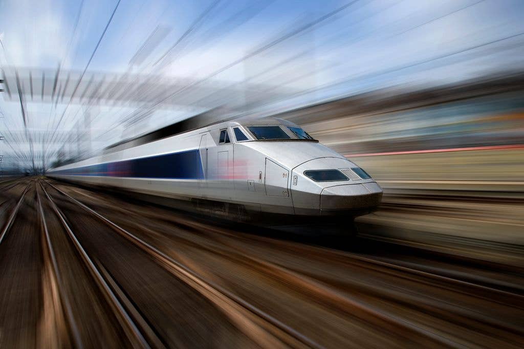 A sleek silver and blue high-speed TGV train speeds along railway tracks, with motion blur emphasising its velocity against a streaked blue sky.
