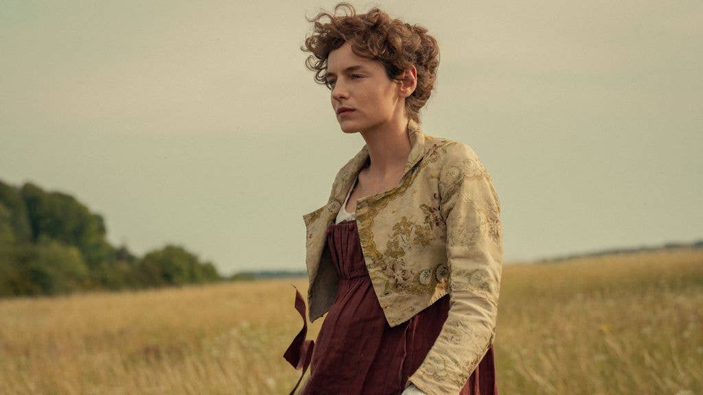 Emma Corrin as Elizabeth Bennet with curly brown hair stands in a field wearing a red dress and a beige jacket.