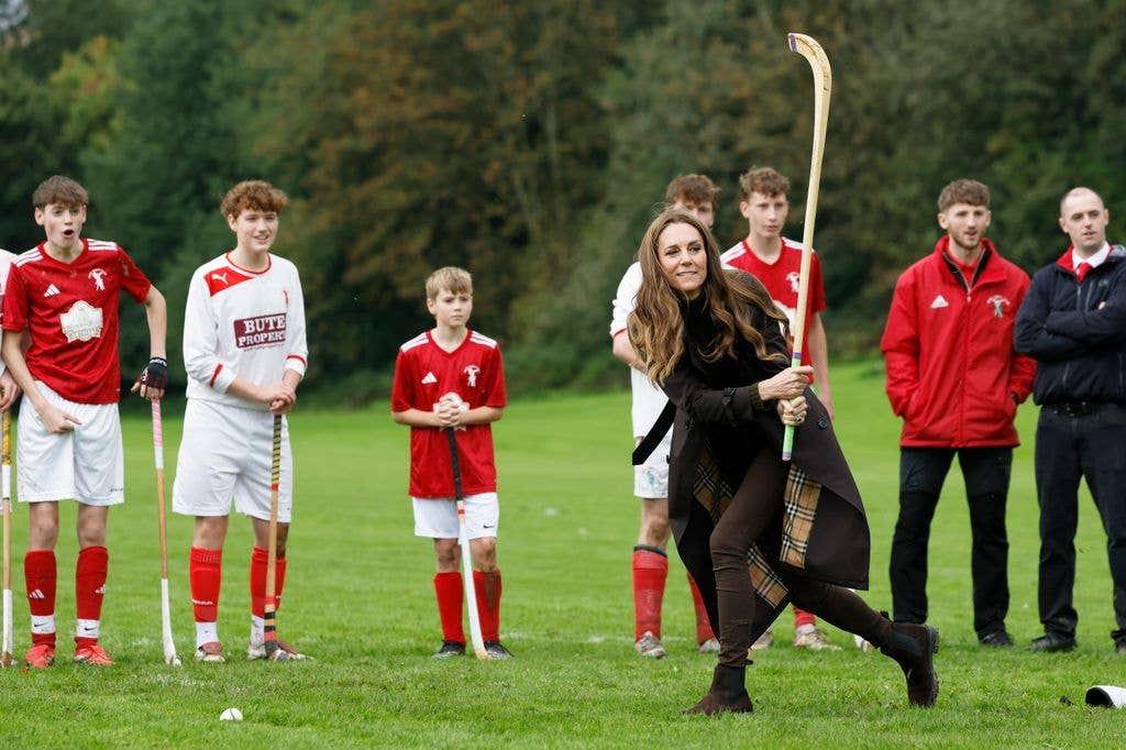 Kate Middleton, in black coat and boots, plays field hockey with young players in red and white uniforms.