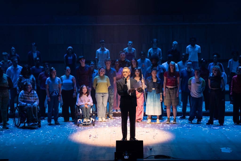 Daniel Craig stands on stage holding a paper, addressing a diverse audience.