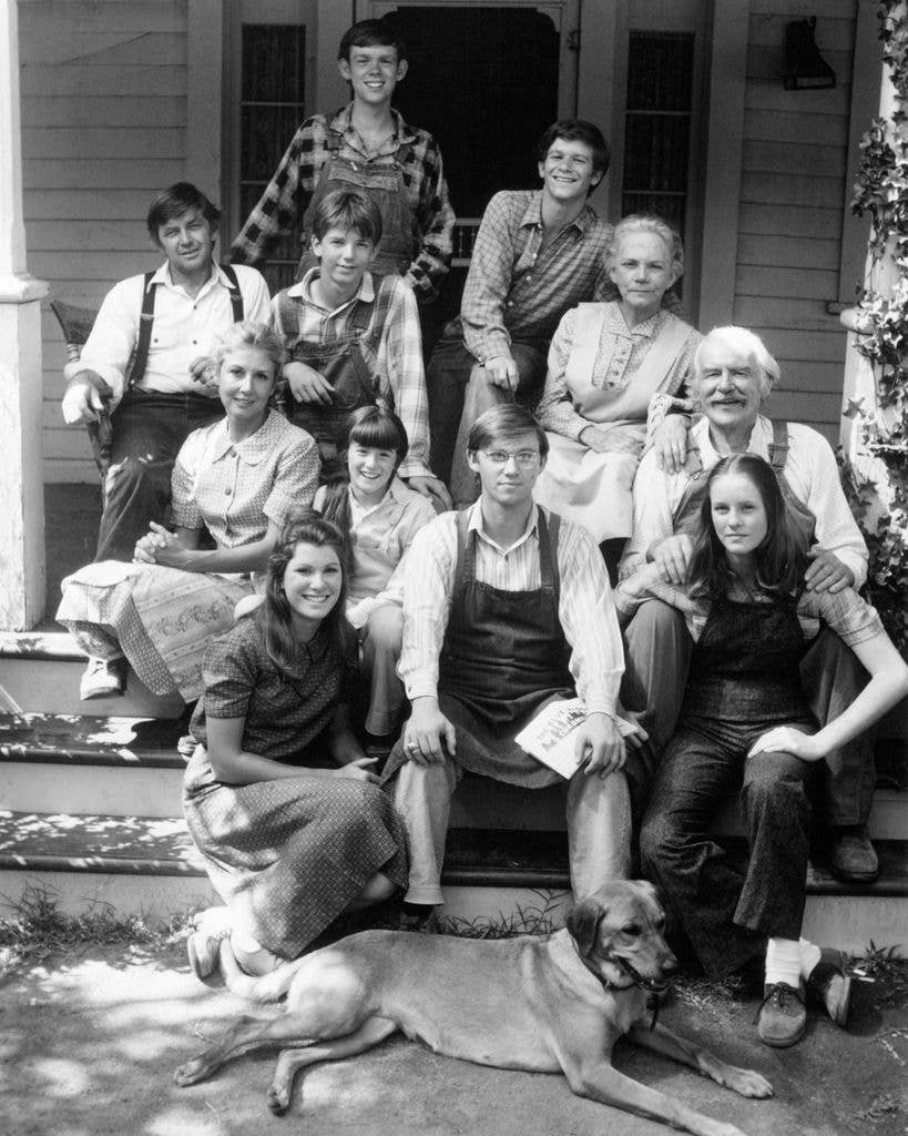 A black and white group portrait of the cast of The Waltons, dressed in period clothing, posed on and around the steps of a wooden porch with a dog lying at the front.