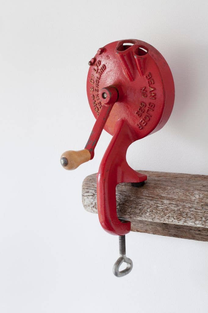 A red enamelled hand-crank bean slicer with a wooden handle grip, clamped by a screw fitting to the edge of an old wooden table against a plain white background.