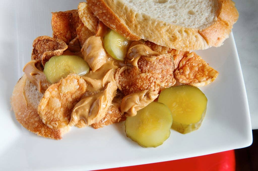 An open sandwich on a white plate is loaded with peanut butter, crisps and sliced pickles.