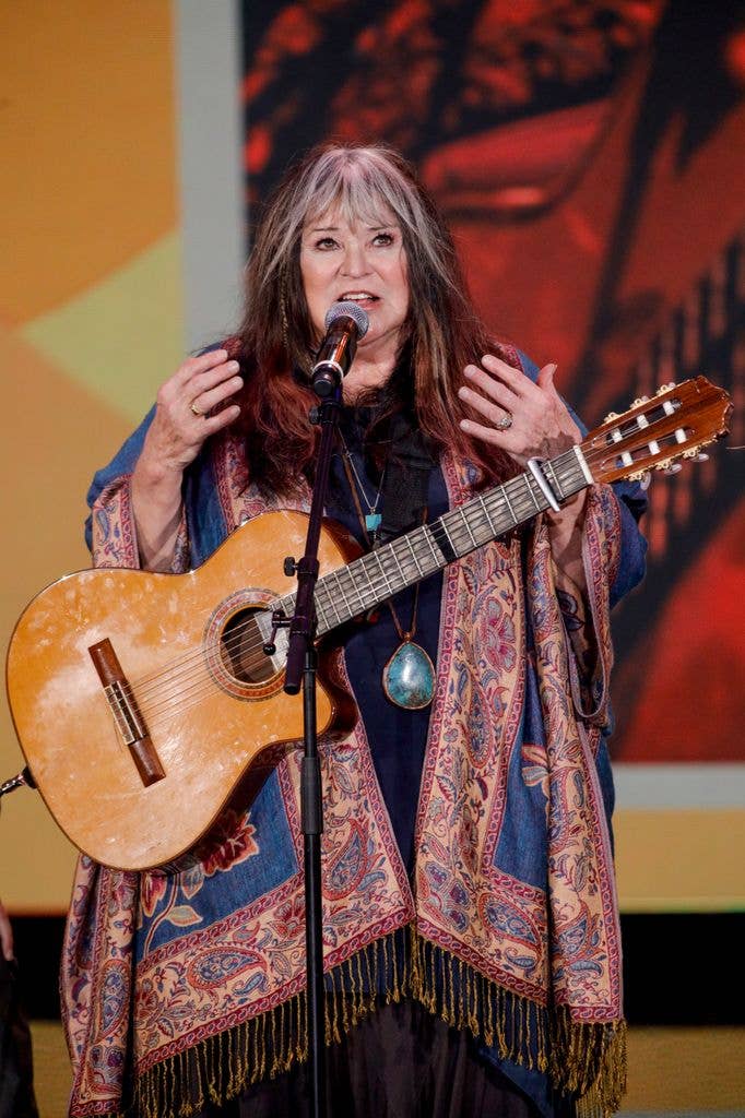 Melanie Safka sings into a microphone with her hands raised, an acoustic guitar slung across her, wearing a paisley shawl.
