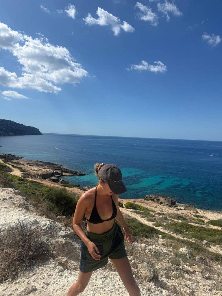 A woman in a black bikini top and green shorts stands on a rocky beach, looking out at the ocean.