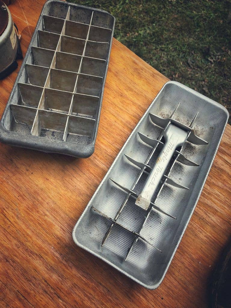 Two old metal ice cube trays sit side by side on a wooden table outdoors, one with a plain grid divider and the other with a lever marked 'Magic Touch' for releasing cubes.
