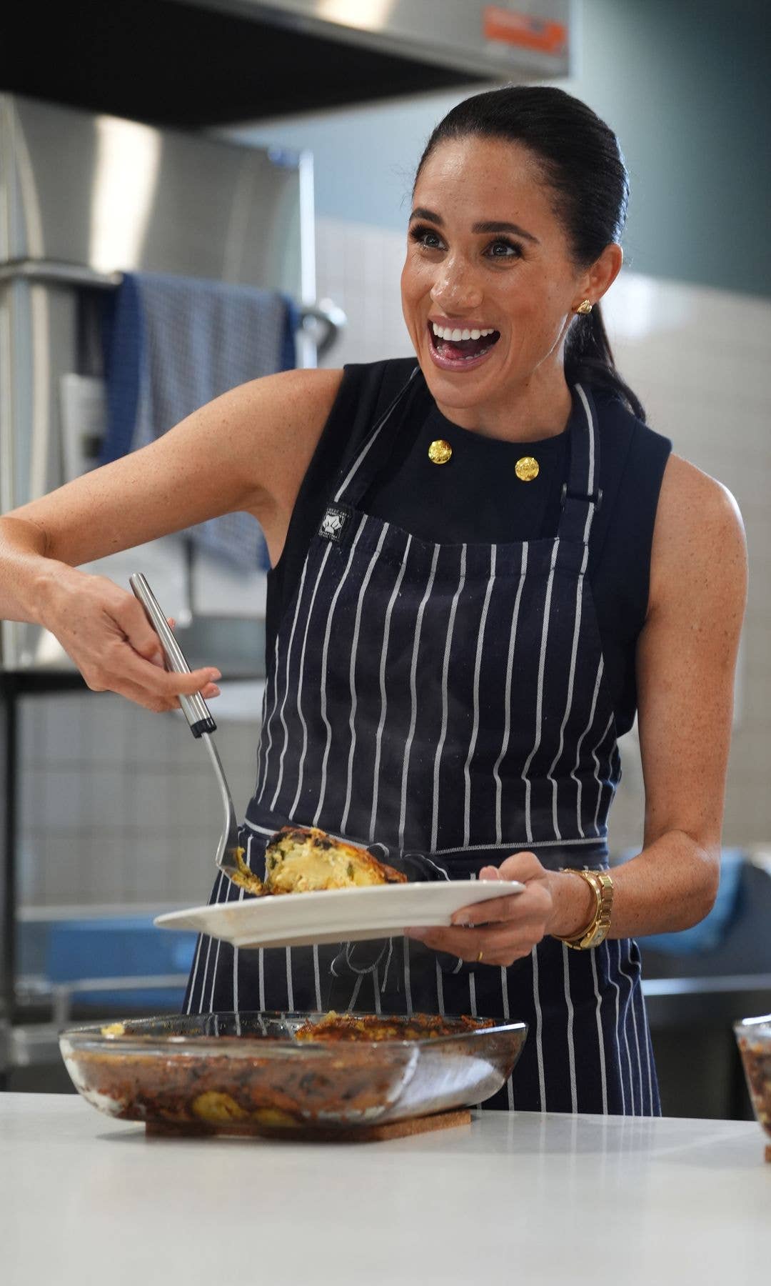 Meghan, Duchess of Sussex, in a black and white striped apron, holding a plate of food with a fork.