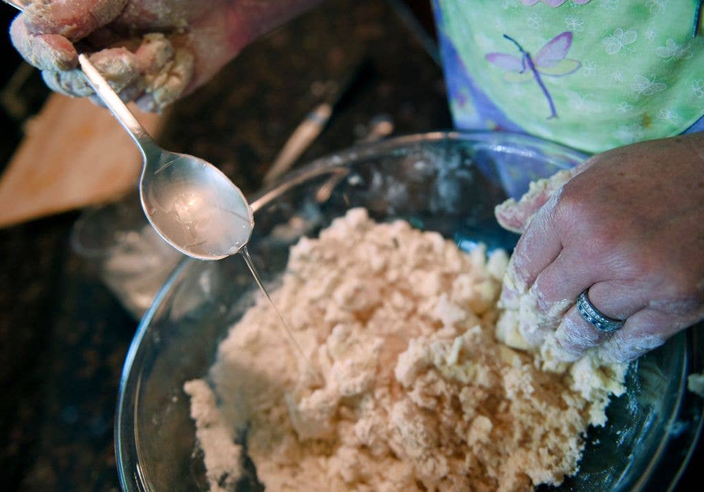 A spoonful of water is drizzled into a bowl of crumbly pastry dough as a hand works the mixture together.