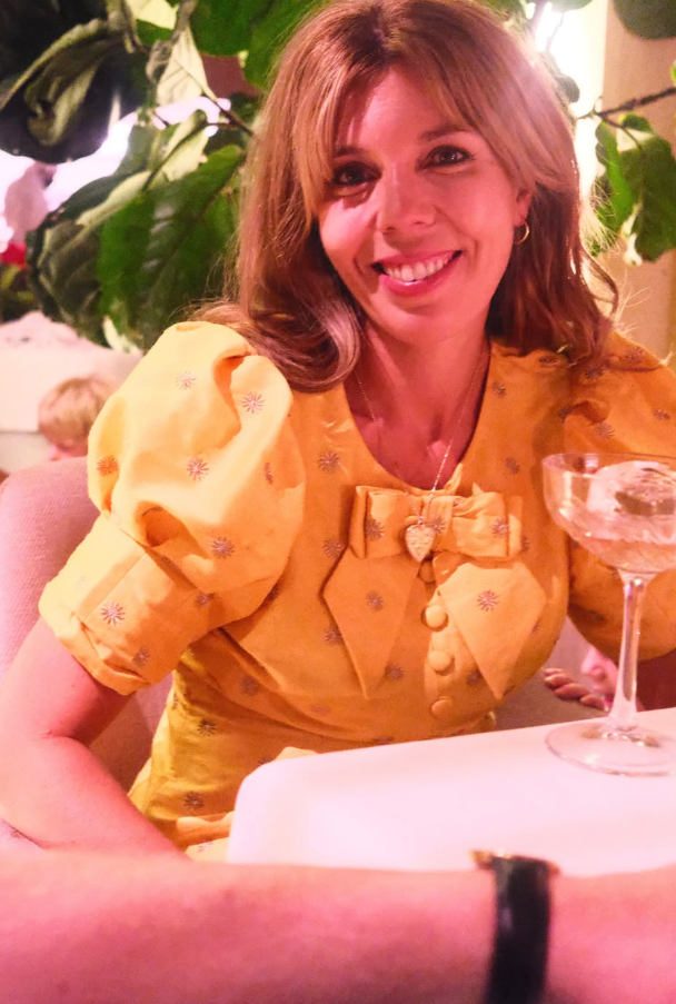 A smiling woman in a yellow dress sits at a table with a wine glass.