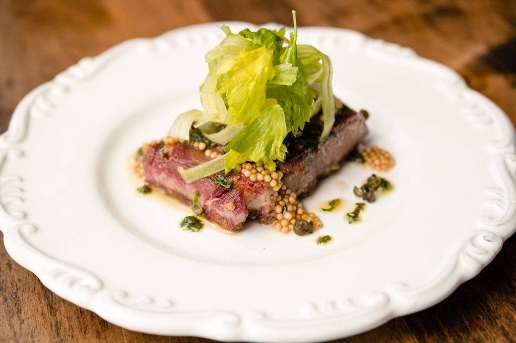 A plated dish of sliced warm beef tongue topped with shaved celery, garnished with capers, herbs and pickled mustard seeds on a white ornate plate.