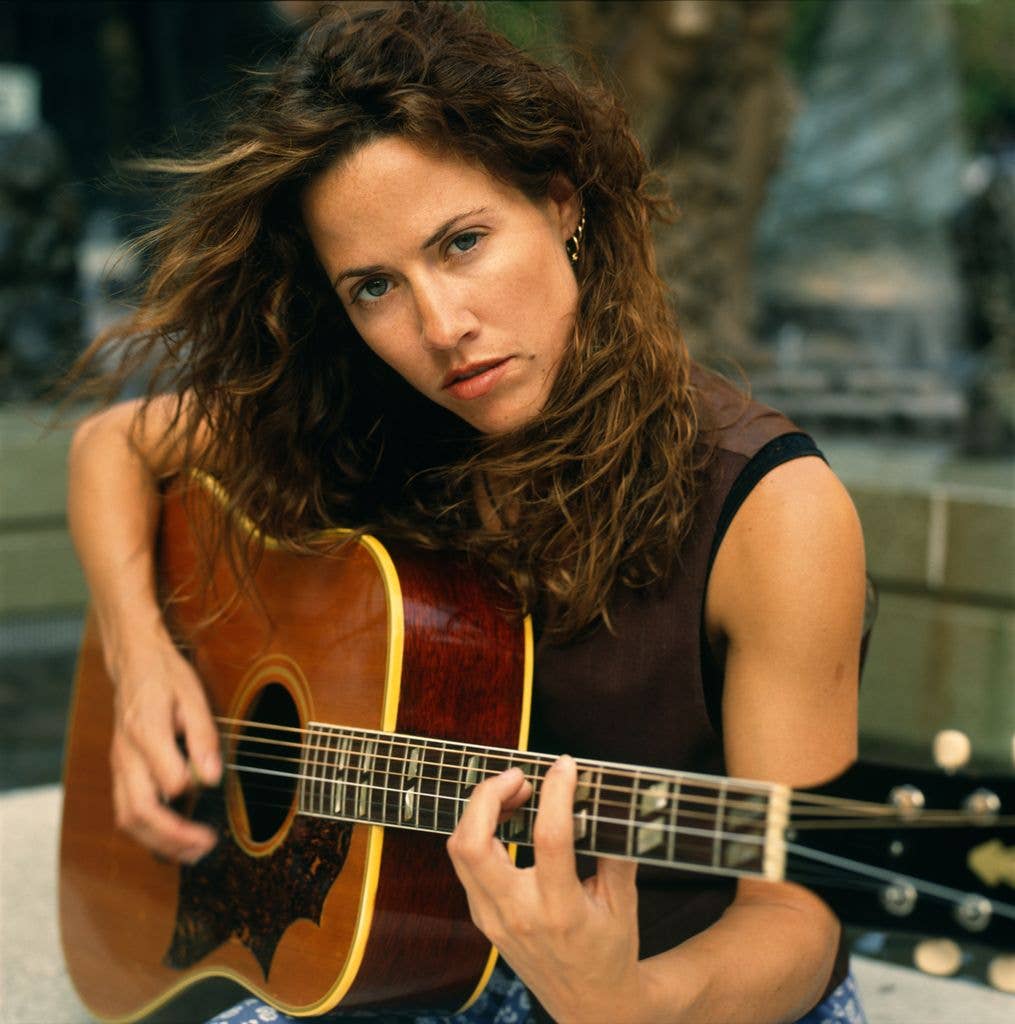 Sheryl Crow, with wind-blown curly hair, looks directly at the camera while holding an acoustic guitar outdoors.
