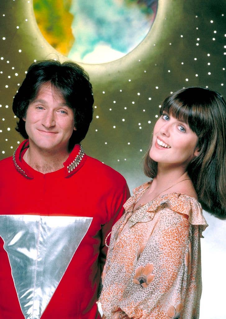 Robin Williams, dressed in a red and silver alien costume, stands smiling beside Pam Dawber in a floral top, both posed against a starry backdrop with a glowing moon.