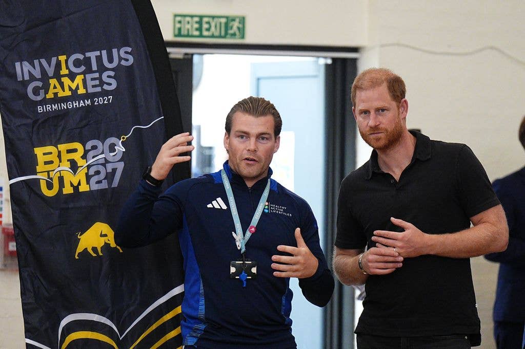 Prince Harry listens to a man wearing a lanyard speaking beside an Invictus Games Birmingham 2027 banner inside a school hall.