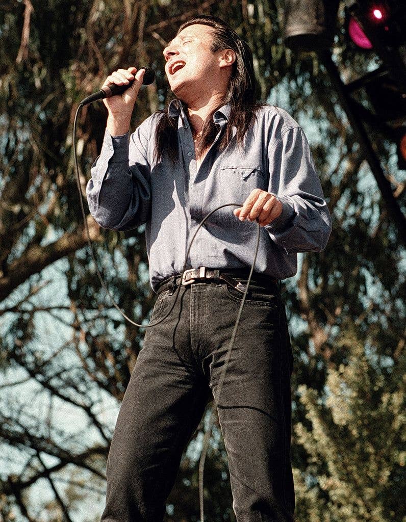 Steve Perry singing on stage, holding a microphone, wearing a blue shirt and black jeans.