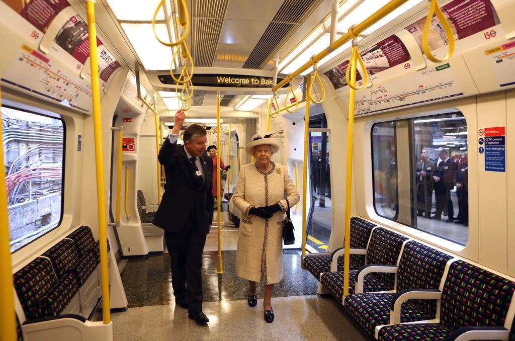 Queen Elizabeth II, wearing a white coat and hat, stands in a train with a man in a suit.
