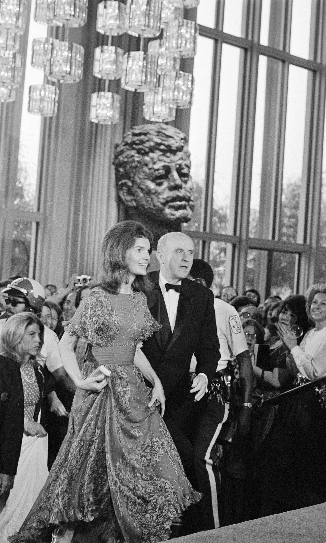 Mrs. Jacqueline Kennedy Onassis passes a huge bronze bust of her late husband, President Kennedy, as she enters the John F. Kennedy Center for the Performing Arts on June 5th. Visiting the Center for the first time, Mrs. Onassis is escorted by Roger Stevens, Chairman of the Board of the Center. The former First Lady attended composer-conductor Leonard Bernstein's production of "Mass," which is dedicated to the late President 1972
