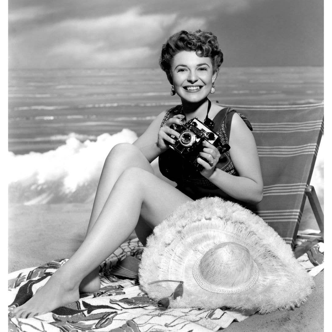 Actress Anne Bancroft (1931-2005) sits by the beach holding an Omega 120 Camera in a publicity shot from 1955, United States. 
