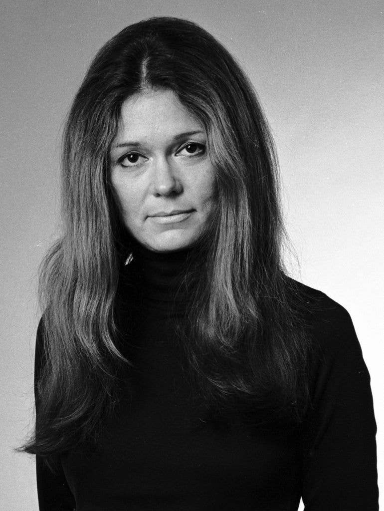 Gloria Steinem, with long hair and wearing a black turtleneck, stands against a plain background.