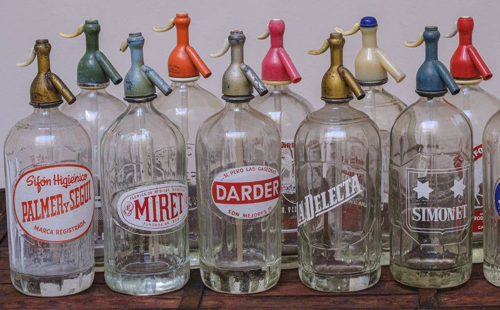A lineup of glass seltzer bottles with metal siphon heads in various colours, labelled with old Spanish brand names including Palmer y Segui, Miret, Darder, La Delecta and Simonet.