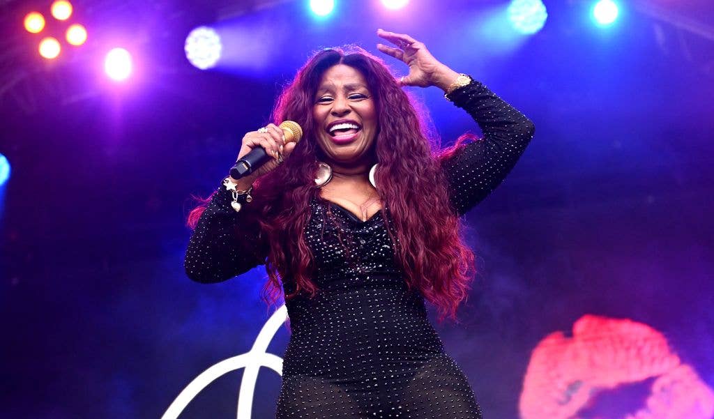 Chaka Khan sings into a microphone with one hand raised, wearing a sparkling black dress under colourful stage lights.