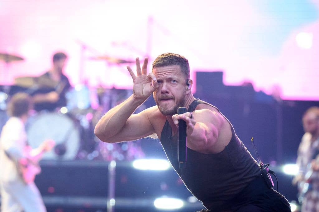 Dan Reynolds of Imagine Dragons leans forward mid-performance, gesturing with one hand near his face while holding a microphone in the other, with bandmates visible on stage behind him.