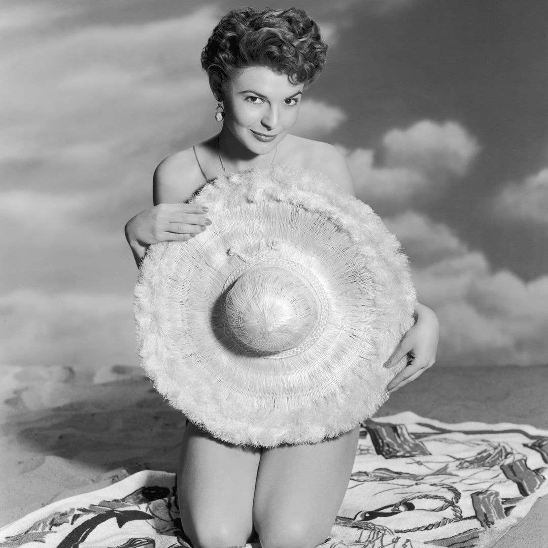 1955:  Studio portrait of American actor Anne Bancroft holding a large straw hat over her nude body while kneeling on a beach towel in sand.