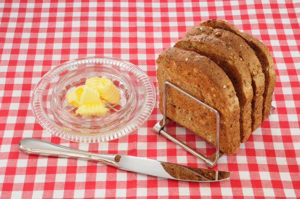 Four slices of seeded wholemeal toast stand in a metal toast rack beside a glass dish of butter curls and a knife on a red and white gingham tablecloth.